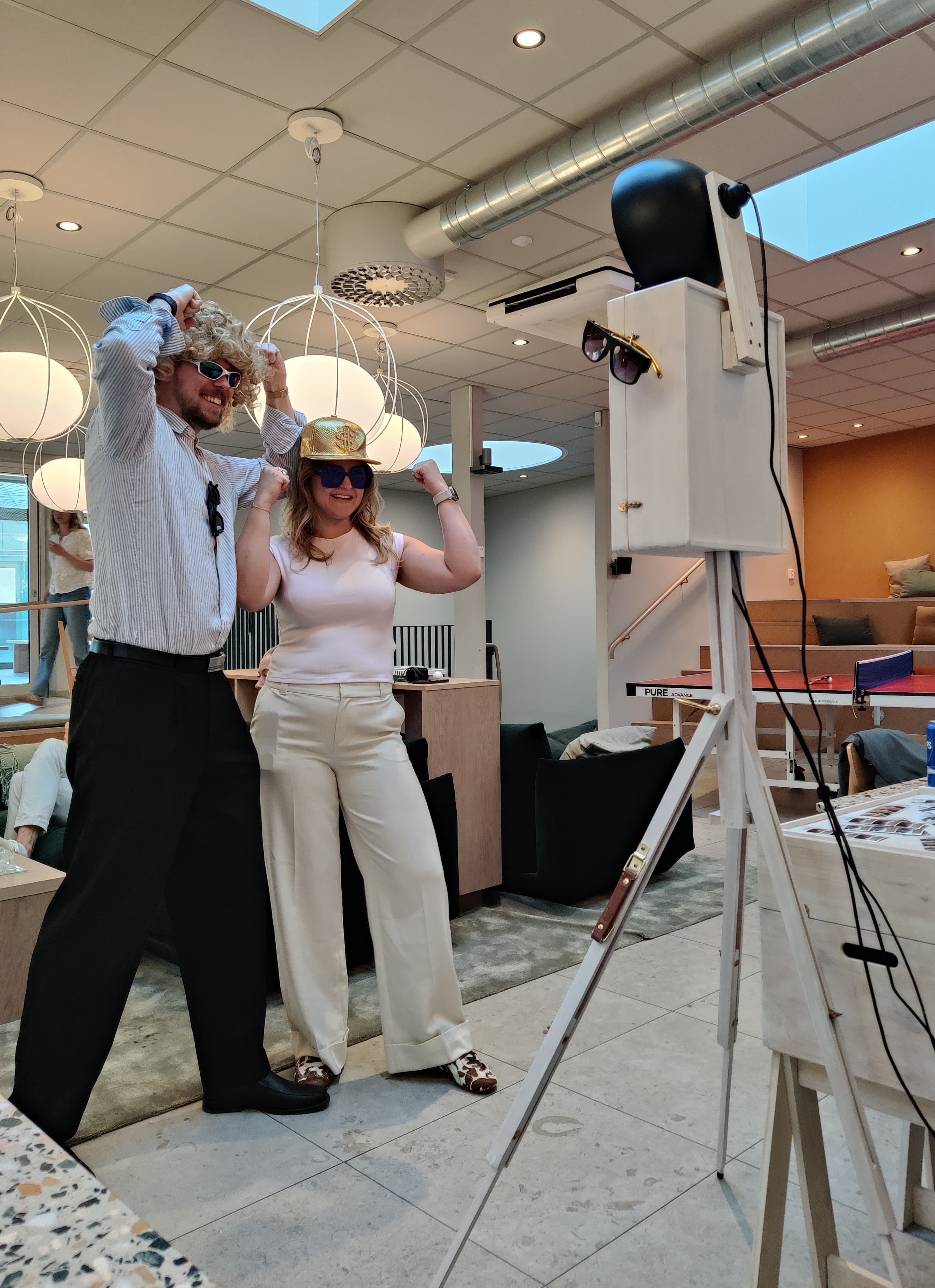 Två kollegor i peruk och guldkeps poserar framför Mabooths fotobås på ett företagsevent